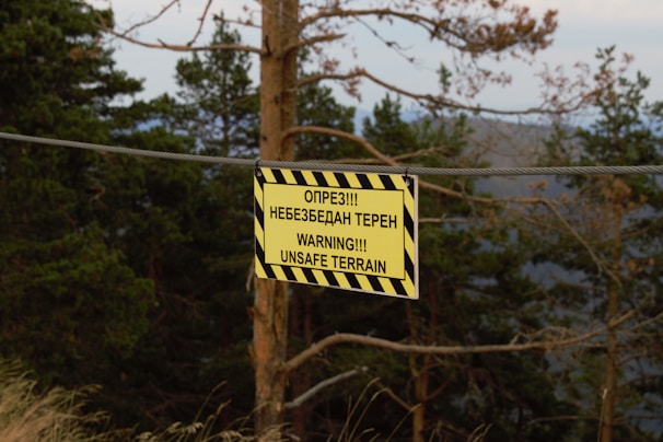 A warning sign hangs from a wire in a forested area. The sign has a yellow background with black diagonal stripes, featuring text in multiple languages warning of unsafe terrain. Trees and foliage are visible in the background, with a slightly overcast sky.