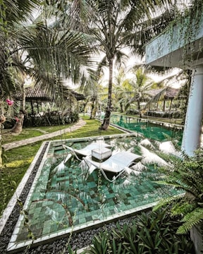 A serene garden scene featuring lush tropical vegetation, including numerous palm trees surrounding a luxuriously tiled water feature. The area includes sun loungers partially submerged in the water, with a path leading to cozy, thatched-roof pavilions in the background. The environment exudes tranquility amidst greenery and water reflections.