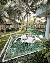 A serene garden scene featuring lush tropical vegetation, including numerous palm trees surrounding a luxuriously tiled water feature. The area includes sun loungers partially submerged in the water, with a path leading to cozy, thatched-roof pavilions in the background. The environment exudes tranquility amidst greenery and water reflections.