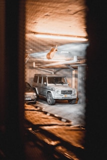 A metallic silver luxury SUV and a smaller black car are parked in an indoor garage. The lighting is warm and dim, casting an amber glow over the concrete ceiling and walls. The vehicles are spotted through a slotted, blurry foreground, suggesting the photo was taken from a distance or through a vent.