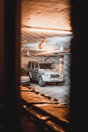 A metallic silver luxury SUV and a smaller black car are parked in an indoor garage. The lighting is warm and dim, casting an amber glow over the concrete ceiling and walls. The vehicles are spotted through a slotted, blurry foreground, suggesting the photo was taken from a distance or through a vent.