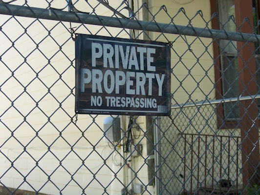 A chain-link fence is prominently featured with a sign that reads 'PRIVATE PROPERTY NO TRESPASSING' attached to it. Behind the fence, there is a building with beige siding and a small railing visible.