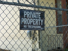 A chain-link fence is prominently featured with a sign that reads 'PRIVATE PROPERTY NO TRESPASSING' attached to it. Behind the fence, there is a building with beige siding and a small railing visible.
