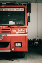 A red fire truck is parked indoors. The vehicle has various labels, including 'FIRE' and a license plate marked 'KA-01 G-8091'. Around the truck, other objects such as helmets and another vehicle can be seen.