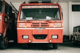 A red fire truck is parked inside a garage. The vehicle has several inscriptions in a local language and English, indicating that it is a TATA fire truck. It is flanked by another similar vehicle on the left and a smaller white vehicle on the right.