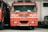 A red fire truck is parked inside a garage. The vehicle has several inscriptions in a local language and English, indicating that it is a TATA fire truck. It is flanked by another similar vehicle on the left and a smaller white vehicle on the right.