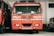 A red fire truck is parked inside a garage. The vehicle has several inscriptions in a local language and English, indicating that it is a TATA fire truck. It is flanked by another similar vehicle on the left and a smaller white vehicle on the right.