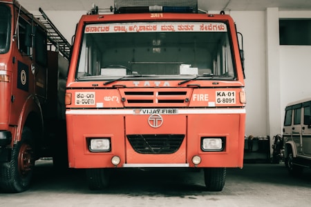 A red fire truck is parked inside a garage. The vehicle has several inscriptions in a local language and English, indicating that it is a TATA fire truck. It is flanked by another similar vehicle on the left and a smaller white vehicle on the right.