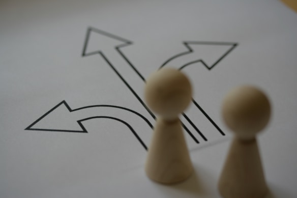 A pair of wooden chess pawns positioned in the foreground, slightly out of focus, with a drawing of several arrows pointing in different directions on a piece of paper in the background.
