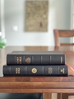 Two black leather-bound Bibles are stacked on a wooden table, with gold lettering indicating their versions: one is the New Revised Standard Version and the other is the English Standard Version. The background includes a blurred image of a wall with an unclear artwork and some green foliage.