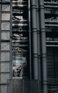 An industrial-style building characterized by large metallic structures and pipes. Visible through a section of glass, three people can be seen engaged in conversation, standing inside what appears to be an elevator or observation area.
