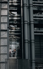 An industrial-style building characterized by large metallic structures and pipes. Visible through a section of glass, three people can be seen engaged in conversation, standing inside what appears to be an elevator or observation area.