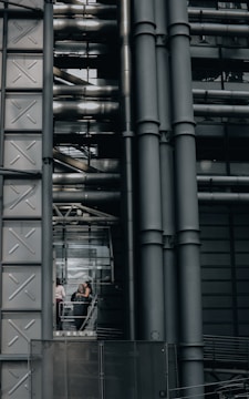An industrial-style building characterized by large metallic structures and pipes. Visible through a section of glass, three people can be seen engaged in conversation, standing inside what appears to be an elevator or observation area.