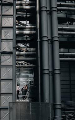 An industrial-style building characterized by large metallic structures and pipes. Visible through a section of glass, three people can be seen engaged in conversation, standing inside what appears to be an elevator or observation area.