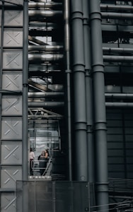 An industrial-style building characterized by large metallic structures and pipes. Visible through a section of glass, three people can be seen engaged in conversation, standing inside what appears to be an elevator or observation area.
