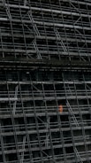 A large, dark building facade under construction, enclosed by an extensive network of scaffolding. One section stands out where a person in a bright orange protective suit is visible.