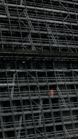 A large, dark building facade under construction, enclosed by an extensive network of scaffolding. One section stands out where a person in a bright orange protective suit is visible.