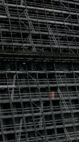 A large, dark building facade under construction, enclosed by an extensive network of scaffolding. One section stands out where a person in a bright orange protective suit is visible.