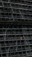 A large, dark building facade under construction, enclosed by an extensive network of scaffolding. One section stands out where a person in a bright orange protective suit is visible.