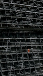 A large, dark building facade under construction, enclosed by an extensive network of scaffolding. One section stands out where a person in a bright orange protective suit is visible.