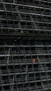 A large, dark building facade under construction, enclosed by an extensive network of scaffolding. One section stands out where a person in a bright orange protective suit is visible.