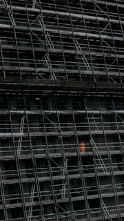 A large, dark building facade under construction, enclosed by an extensive network of scaffolding. One section stands out where a person in a bright orange protective suit is visible.