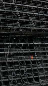 A large, dark building facade under construction, enclosed by an extensive network of scaffolding. One section stands out where a person in a bright orange protective suit is visible.