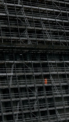 A large, dark building facade under construction, enclosed by an extensive network of scaffolding. One section stands out where a person in a bright orange protective suit is visible.