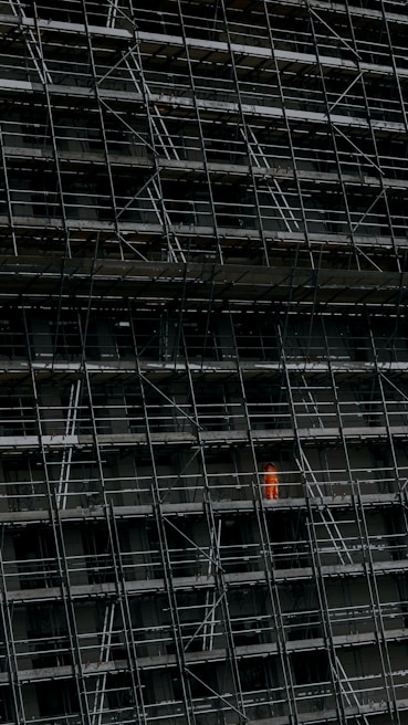 A large, dark building facade under construction, enclosed by an extensive network of scaffolding. One section stands out where a person in a bright orange protective suit is visible.