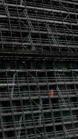A large, dark building facade under construction, enclosed by an extensive network of scaffolding. One section stands out where a person in a bright orange protective suit is visible.