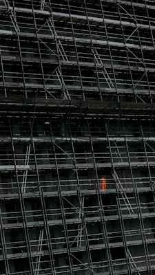 A large, dark building facade under construction, enclosed by an extensive network of scaffolding. One section stands out where a person in a bright orange protective suit is visible.