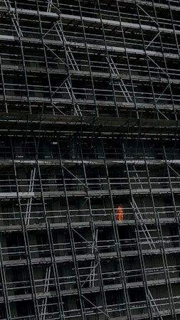 A large, dark building facade under construction, enclosed by an extensive network of scaffolding. One section stands out where a person in a bright orange protective suit is visible.