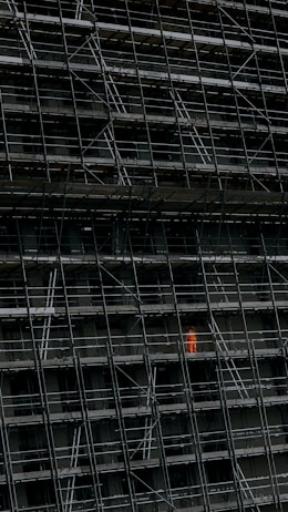 A large, dark building facade under construction, enclosed by an extensive network of scaffolding. One section stands out where a person in a bright orange protective suit is visible.