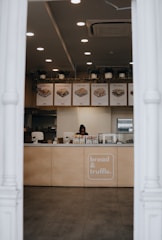 A sandwich shop with a counter displaying various food items and a person working. The menu items displayed above the counter include different types of sandwiches. The shop has a modern interior with several ceiling lights, an air conditioning unit, and potted plants hanging above the counter.