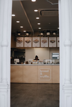 A sandwich shop with a counter displaying various food items and a person working. The menu items displayed above the counter include different types of sandwiches. The shop has a modern interior with several ceiling lights, an air conditioning unit, and potted plants hanging above the counter.