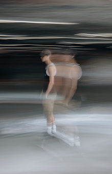 A blurred motion capture of a person on a skateboard, creating a ghostly effect. The person appears to be moving, with their image duplicated and smeared across the scene. The background is dark and indistinct, emphasizing the movement of the figure.