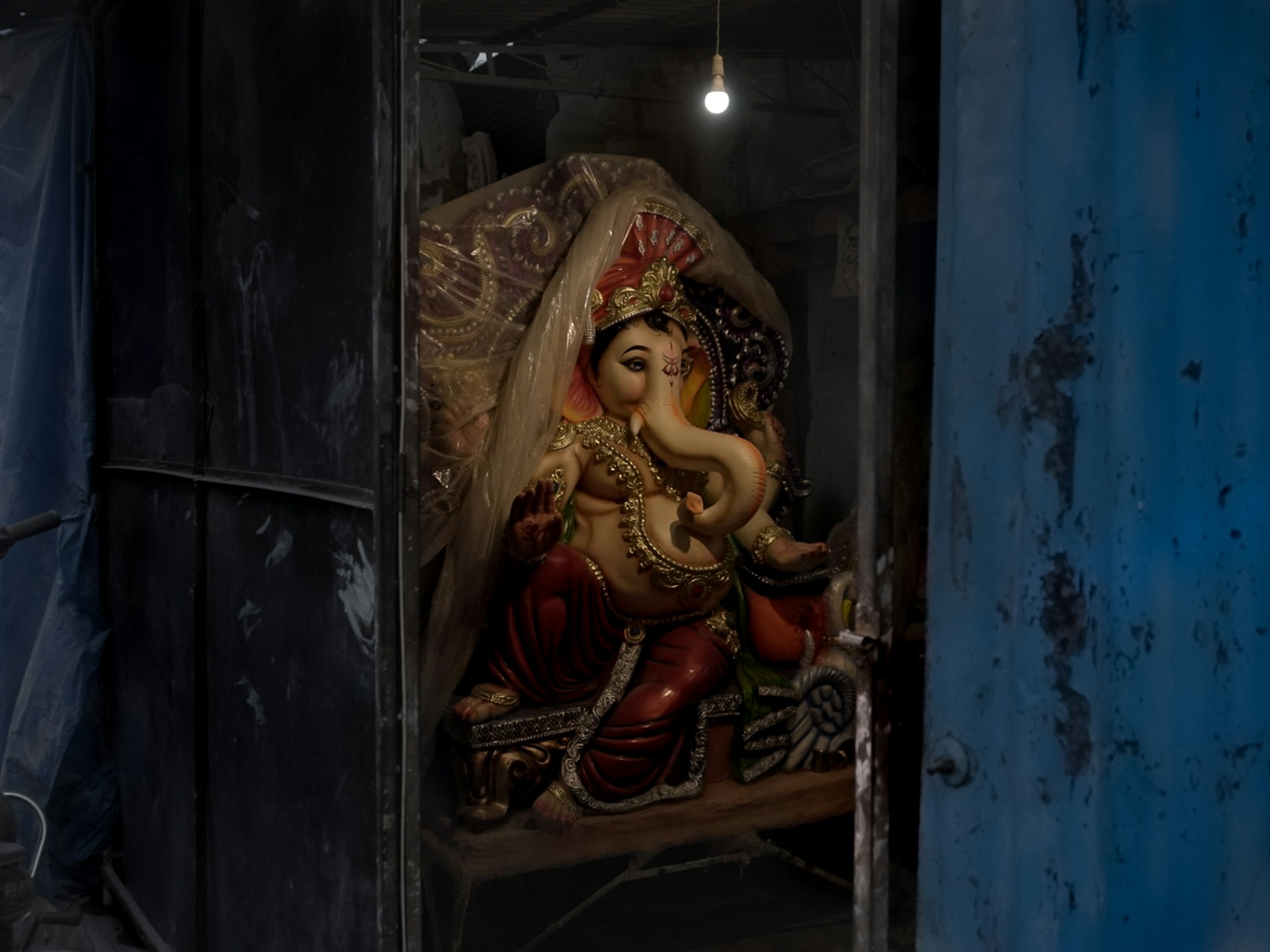A statue of Lord Ganesha is placed inside a dimly lit room, partially covered by a transparent plastic sheet with a glowing light bulb hanging above. The statue is intricately decorated, showcasing detailed ornaments and vibrant colors.