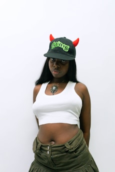 A person with long dark hair wears a white crop top, olive green pants, and a black baseball cap adorned with red devil horns and the word 'Saints' in green text. A large pendant necklace is visible.