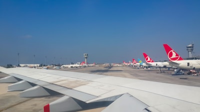 An airport scene with multiple aircraft lined up on the tarmac, featuring red and white tail fin designs. A prominent airplane wing is visible in the foreground. A control tower is positioned in the background under a clear blue sky.