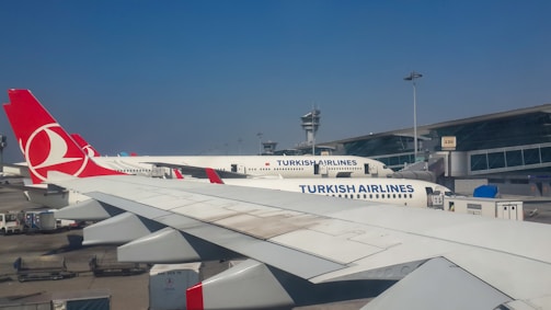Several Turkish Airlines planes are parked at an airport, with a focus on the aircraft's wings and tailfins showing the airline's logo. The airport terminal and control tower are visible in the background under a clear blue sky.