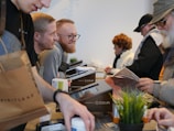 A bustling scene with several people engaged in transactions or interactions across a counter. A variety of items like a brown shopping bag, documents, and a potted plant are visible on the countertop. Individuals appear to be wearing casual attire, including sweaters and hats, suggesting an informal setting.