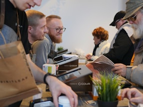 A bustling scene with several people engaged in transactions or interactions across a counter. A variety of items like a brown shopping bag, documents, and a potted plant are visible on the countertop. Individuals appear to be wearing casual attire, including sweaters and hats, suggesting an informal setting.