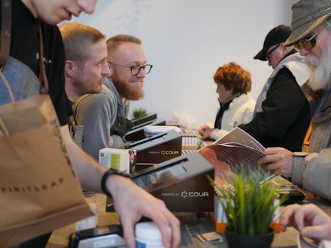 A bustling scene with several people engaged in transactions or interactions across a counter. A variety of items like a brown shopping bag, documents, and a potted plant are visible on the countertop. Individuals appear to be wearing casual attire, including sweaters and hats, suggesting an informal setting.