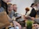 A bustling scene with several people engaged in transactions or interactions across a counter. A variety of items like a brown shopping bag, documents, and a potted plant are visible on the countertop. Individuals appear to be wearing casual attire, including sweaters and hats, suggesting an informal setting.