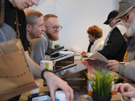 A bustling scene with several people engaged in transactions or interactions across a counter. A variety of items like a brown shopping bag, documents, and a potted plant are visible on the countertop. Individuals appear to be wearing casual attire, including sweaters and hats, suggesting an informal setting.