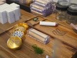 A wooden tray holds various smoking-related items, including a metallic grinder with a gold finish, a white and red lighter, pre-rolled cones in a holder, and some green herbs scattered nearby. There are also small glass jars and packaged boxes in the background.