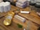 A wooden tray holds various smoking-related items, including a metallic grinder with a gold finish, a white and red lighter, pre-rolled cones in a holder, and some green herbs scattered nearby. There are also small glass jars and packaged boxes in the background.
