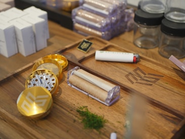 A wooden tray holds various smoking-related items, including a metallic grinder with a gold finish, a white and red lighter, pre-rolled cones in a holder, and some green herbs scattered nearby. There are also small glass jars and packaged boxes in the background.