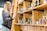 A person stands beside a wooden shelving unit holding various jars and containers. They are observing the contents of the shelves while holding a tablet. The setting appears to be a store or an apothecary styled space with warm, wooden interiors.