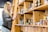 A person stands beside a wooden shelving unit holding various jars and containers. They are observing the contents of the shelves while holding a tablet. The setting appears to be a store or an apothecary styled space with warm, wooden interiors.